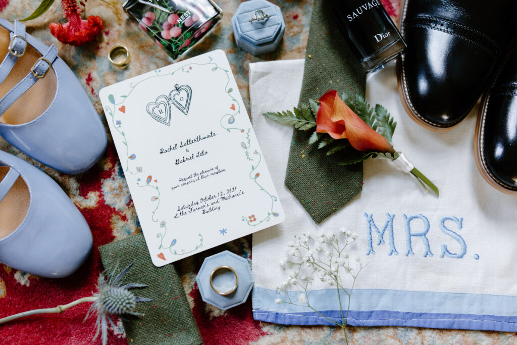 Flat lay of wedding day details including blue shoes, invite suite, groom's green tie and florals.